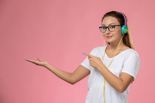 Vista frontal joven mujer atractiva en camiseta blanca posando y escuchando música a través de auriculares sonriendo sobre el fondo rosa