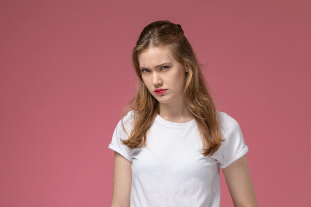 Vista frontal joven mujer atractiva en camiseta blanca posando con expresión de disgusto en la pared rosa modelo pose femenina fotografía en color