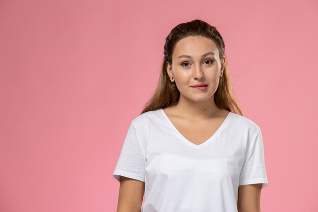 Vista frontal joven mujer atractiva en camiseta blanca posando con expresión linda sobre fondo rosa