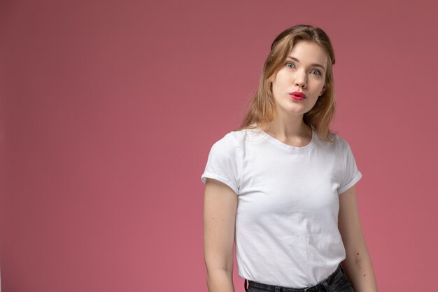 Vista frontal joven mujer atractiva en camiseta blanca posando con expresión de sorpresa en la pared rosa modelo pose femenina color hembra joven