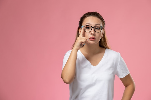 Vista frontal joven mujer atractiva en camiseta blanca posando con gafas de sol ópticas en el fondo rosa