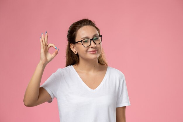 Vista frontal joven mujer atractiva en camiseta blanca posando y guiñando un ojo smi sobre fondo rosa