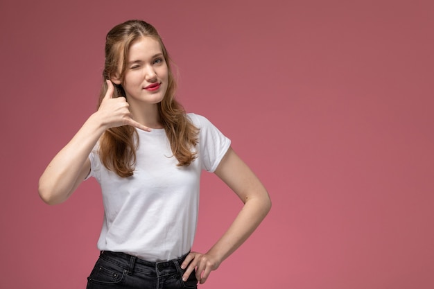 Vista frontal joven mujer atractiva en camiseta blanca posando guiñando un ojo y sonriendo en la pared rosa modelo pose femenina fotografía en color