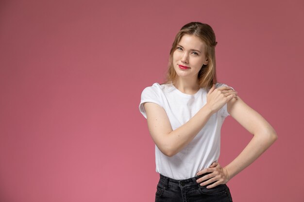 Vista frontal joven mujer atractiva en camiseta blanca posando con una leve sonrisa en la pared rosa modelo color mujer joven