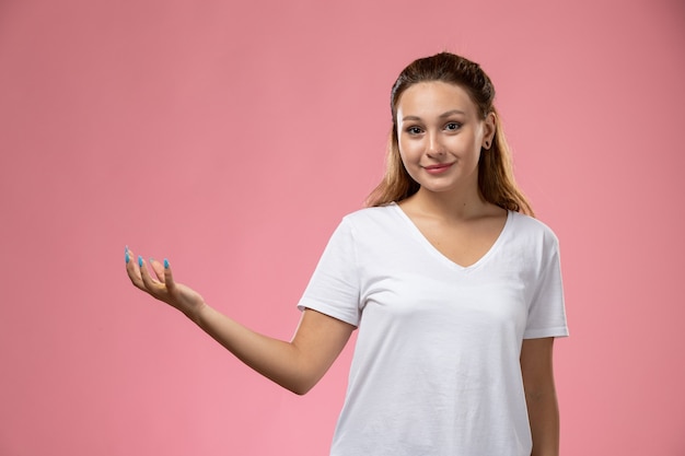 Vista frontal joven mujer atractiva en camiseta blanca posando con una ligera sonrisa sobre fondo rosa