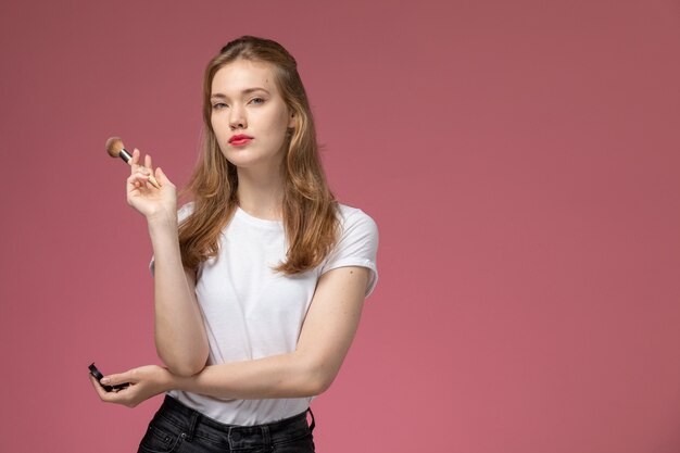 Vista frontal joven mujer atractiva en camiseta blanca posando con pequeño espejo y pincel en la pared rosa modelo color mujer joven