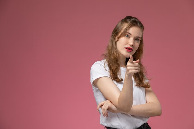 Vista frontal joven mujer atractiva en camiseta blanca posando señalando en la pared rosa modelo pose femenina fotografía en color