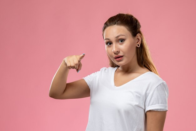 Vista frontal joven mujer atractiva en camiseta blanca posando señalando sobre el fondo rosa