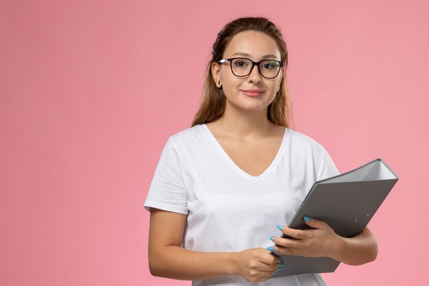 Vista frontal joven mujer atractiva en camiseta blanca posando y smi sosteniendo un archivo gris en el escritorio rosa