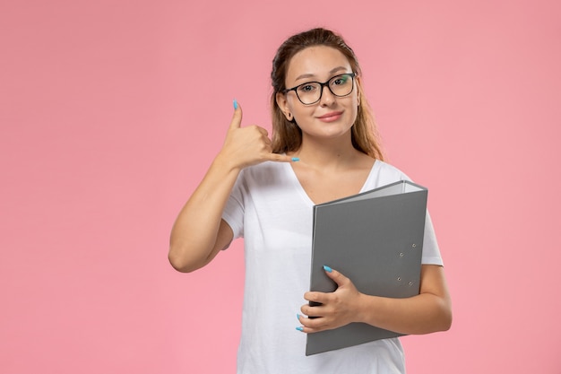 Vista frontal joven mujer atractiva en camiseta blanca posando y smi sosteniendo un archivo gris sobre fondo rosa