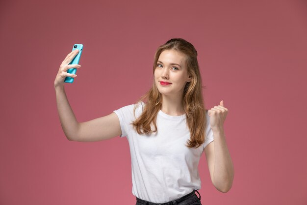 Vista frontal joven mujer atractiva en camiseta blanca posando sonriendo y tomando un selfie en pared rosa modelo pose femenina foto en color joven mujer