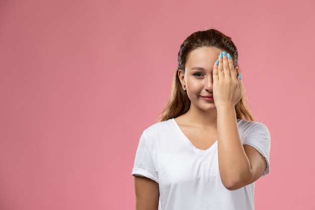 Vista frontal joven mujer atractiva en camiseta blanca posando con una sonrisa y cubriendo un lado de su rostro sobre fondo rosa