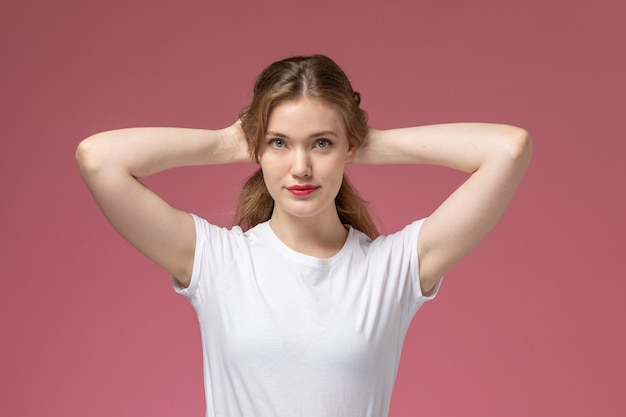 Vista frontal joven mujer atractiva en camiseta blanca posando sosteniendo su cabello en la pared rosa modelo pose femenina color hembra joven