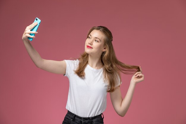 Vista frontal joven mujer atractiva en camiseta blanca posando y tomando un selfie en la pared rosa modelo color mujer joven