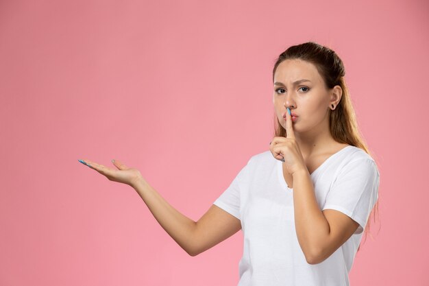 Vista frontal joven mujer atractiva en camiseta blanca que muestra el signo de silencio sobre el fondo rosa