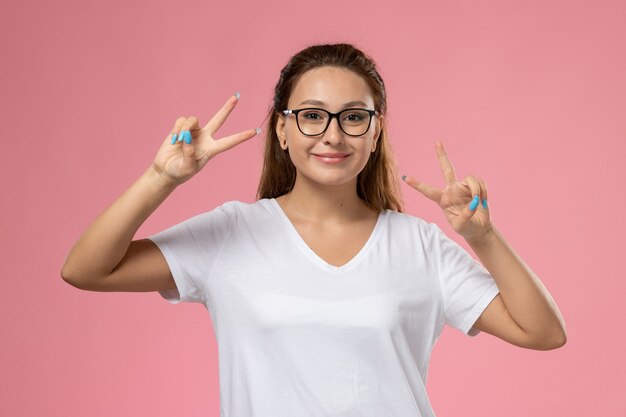 Vista frontal joven mujer atractiva en camiseta blanca smi con los dedos levantados gesto de victoria sobre el fondo rosa