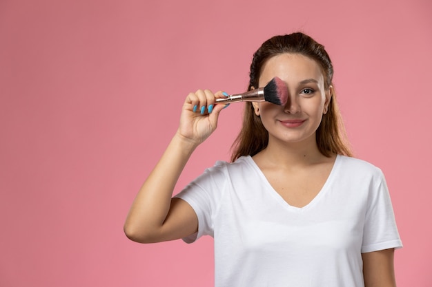 Vista frontal joven mujer atractiva en camiseta blanca smi y sosteniendo pincel de maquillaje sobre fondo rosa