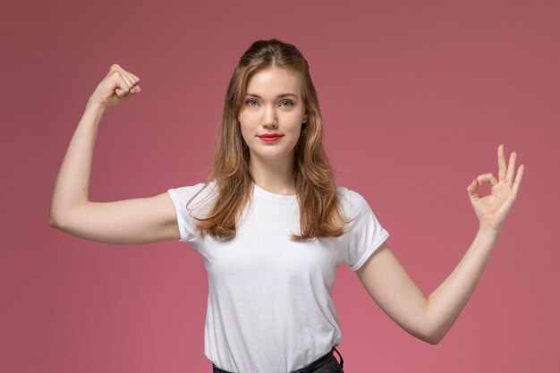 Vista frontal joven mujer atractiva en camiseta blanca sonriendo flexionando sobre la pared de color rosa oscuro modelo color mujer joven