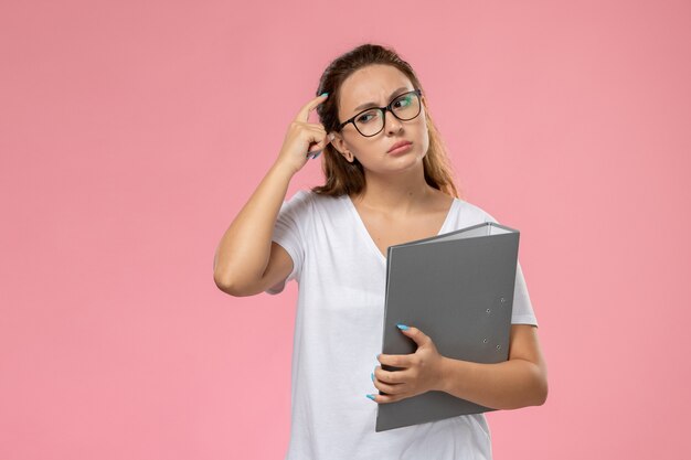 Vista frontal joven mujer atractiva en camiseta blanca sosteniendo un archivo gris y pensando en el fondo rosa