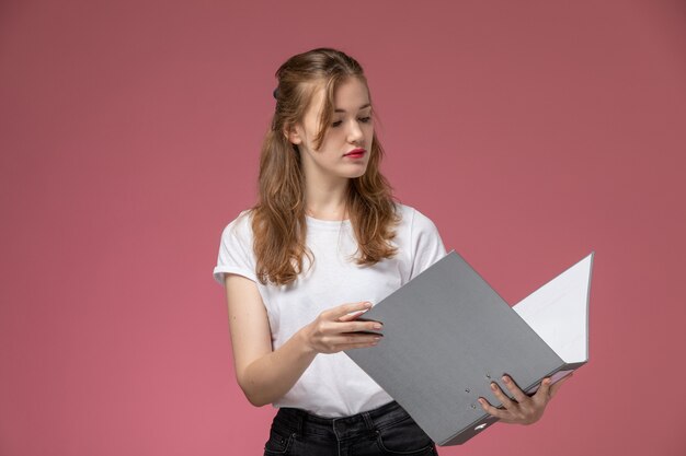 Vista frontal joven mujer atractiva en camiseta blanca sosteniendo documento gris leyéndolo en la pared rosa modelo pose femenina color hembra joven