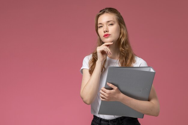 Vista frontal joven mujer atractiva en camiseta blanca sosteniendo documento gris y pensando en la pared rosa modelo pose femenina color hembra joven