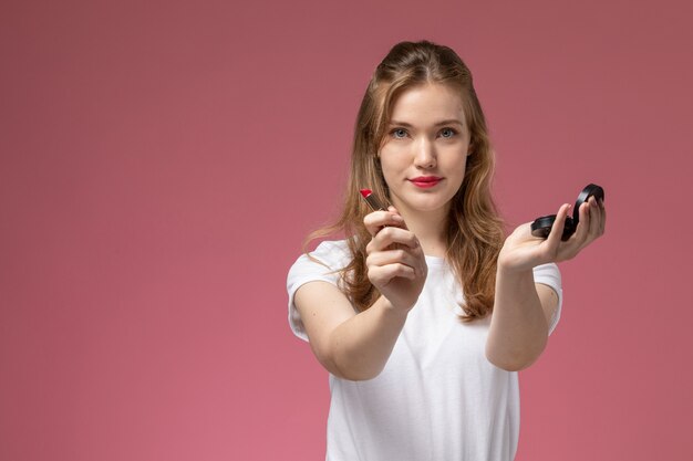 Vista frontal joven mujer atractiva en camiseta blanca sosteniendo lápiz labial con sonrisa en la pared rosa modelo color mujer joven