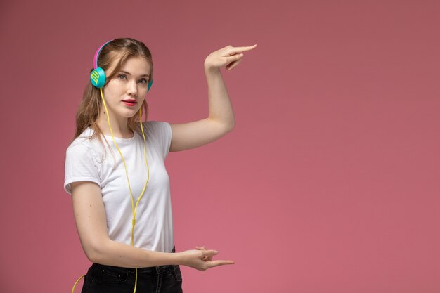 Vista frontal joven mujer atractiva en camiseta blanca usando sus auriculares y mostrando el tamaño con sus manos en la pared de color rosa oscuro modelo color mujer joven