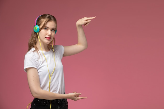 Vista frontal joven mujer atractiva en camiseta blanca usando sus auriculares y mostrando el tamaño con sus manos en la pared de color rosa oscuro modelo color mujer joven