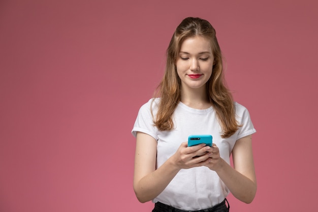 Vista frontal joven mujer atractiva en camiseta blanca usando un teléfono con una leve sonrisa en la pared rosa modelo pose femenina fotografía en color