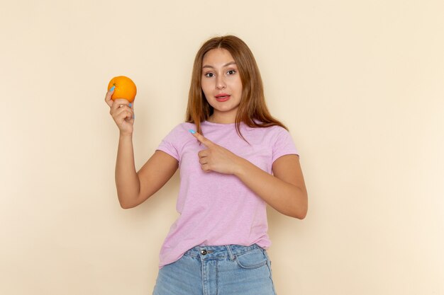 Vista frontal joven mujer atractiva en camiseta rosa y jeans sosteniendo y apuntando la naranja