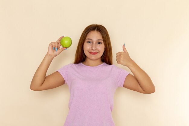 Vista frontal joven mujer atractiva en camiseta rosa y jeans sosteniendo manzana y posando con sonrisa