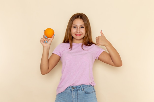 Vista frontal joven mujer atractiva en camiseta rosa y jeans sosteniendo naranja y mostrando un signo impresionante