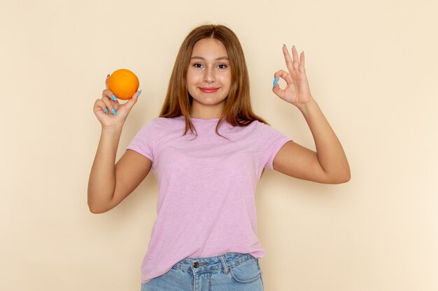 Vista frontal joven mujer atractiva en camiseta rosa y jeans sosteniendo naranja y sonriendo