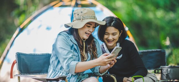 Vista frontal Joven mujer bonita asiática y su novia sentadas frente a la tienda usan el teléfono móvil para tomar una foto durante el campamento en el bosque con felicidad juntos