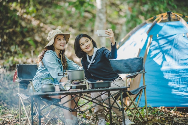 Vista frontal Joven mujer bonita asiática y su novia sentadas frente a la tienda usan el teléfono móvil para tomar una foto durante el campamento en el bosque con felicidad juntos