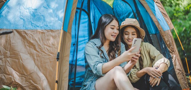 Vista frontal Joven mujer bonita asiática y su novia sentadas frente a la tienda usan el teléfono móvil para tomar una foto durante el campamento en el bosque con felicidad juntos