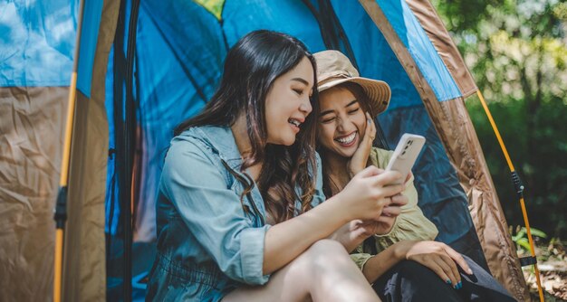 Vista frontal Joven mujer bonita asiática y su novia sentadas frente a la tienda usan el teléfono móvil para tomar una foto durante el campamento en el bosque con felicidad juntos