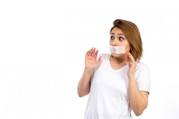 Una vista frontal joven mujer en camiseta blanca con una venda blanca alrededor de la boca por miedo a las amenazas en el blanco