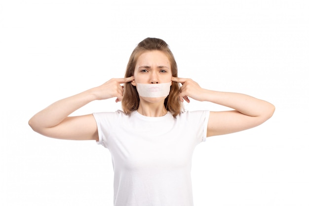 Una vista frontal joven mujer en camiseta blanca con vendaje blanco alrededor de su boca cerrando sus oídos en el blanco
