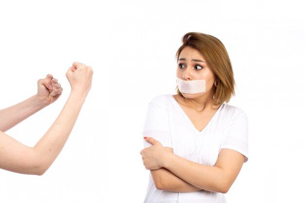 Una vista frontal joven mujer en camiseta blanca con vendaje blanco alrededor de su boca se declara culpable de miedo expresión en el blanco