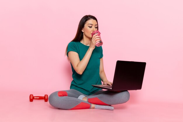 Vista frontal joven mujer en camiseta verde usando su computadora portátil en la pared rosa claro cintura deporte ejercicio entrenamientos belleza delgada atleta femenina