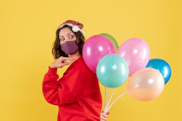 Vista frontal joven mujer escondiendo globos en máscara estéril sobre un fondo amarillo color vacaciones emoción año nuevo mujer de navidad