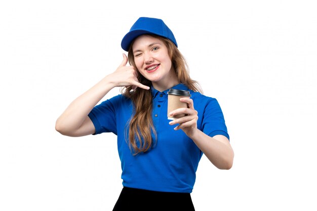 Una vista frontal joven mujer mensajero en camisa azul gorra azul sonriendo mostrando señal de llamada telefónica sosteniendo una taza de café en blanco