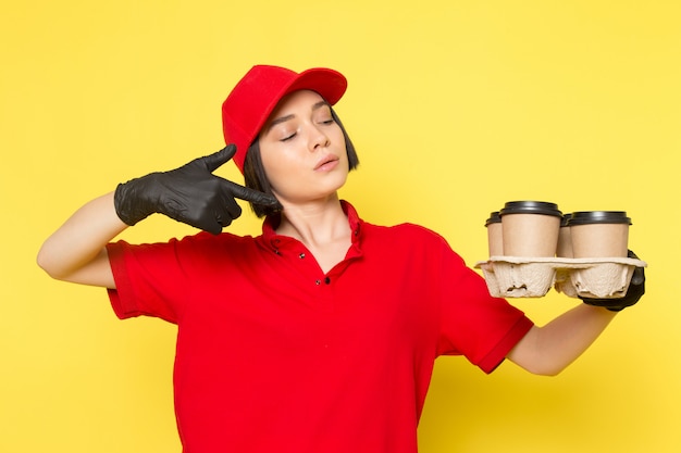 Una vista frontal joven mujer mensajero en rojo uniforme guantes negros y gorra roja con tazas de café