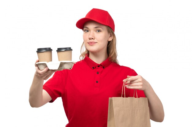 Una vista frontal joven mujer mensajero trabajadora del servicio de entrega de alimentos sonriendo sosteniendo el paquete de entrega y tazas con café en blanco