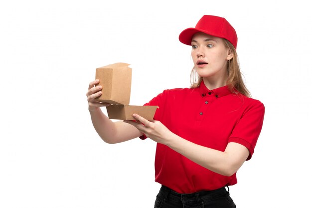 Una vista frontal joven mujer mensajero trabajadora del servicio de entrega de alimentos sosteniendo y abriendo el paquete de entrega de alimentos con expresión de sorpresa en blanco