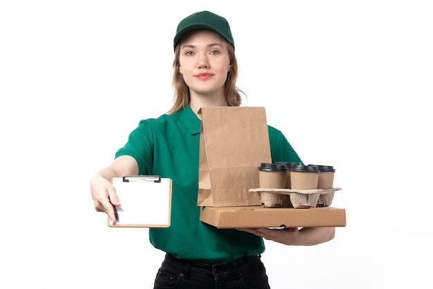 Una vista frontal joven mujer mensajero en uniforme verde sosteniendo paquetes de taza de café bloc de notas y sonriendo en blanco