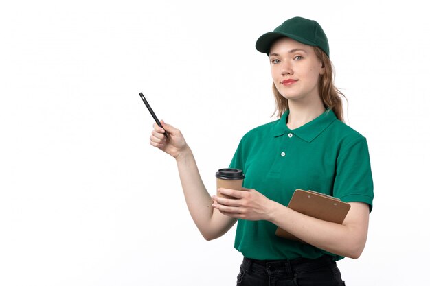 Una vista frontal joven mujer mensajero en uniforme verde sosteniendo la taza de café y el bloc de notas sonriendo en blanco