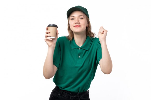 Una vista frontal joven mujer mensajero en uniforme verde sosteniendo la taza de café y sonriendo en blanco