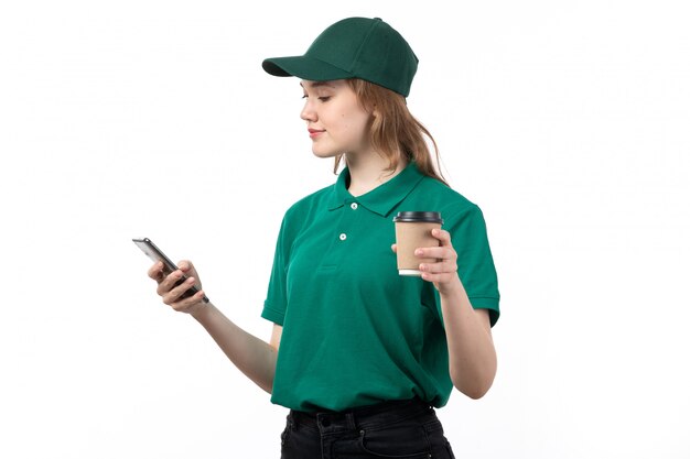 Una vista frontal joven mujer mensajero en uniforme verde sosteniendo la taza de café y usando un teléfono en blanco